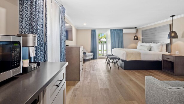 A modern hotel room with a kitchenette, a bed with a grey headboard, a grey sofa, and a view of the outdoors through a sliding door. - Love Farms Lodge (West Dennis)