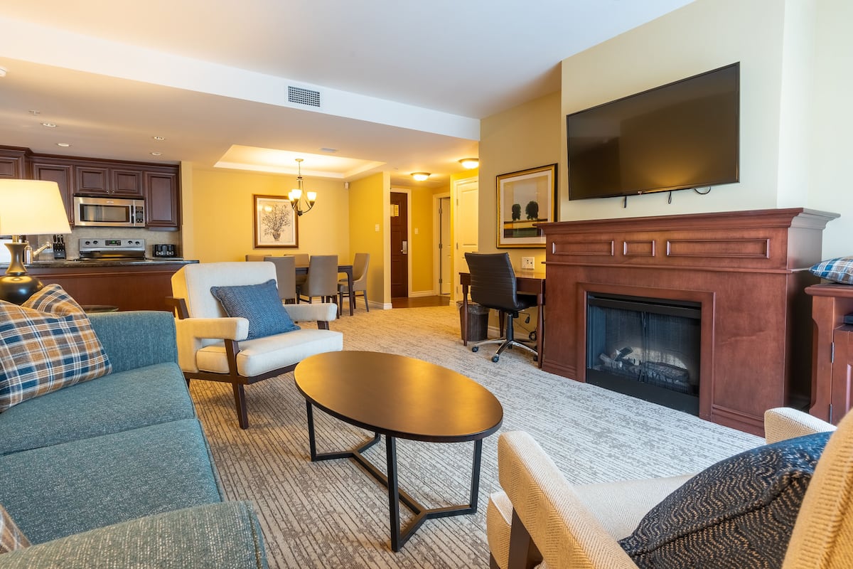 A living room with a sofa, armchairs, a coffee table, a fireplace, a television, and a kitchen area with a microwave.
