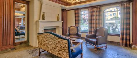 A spacious room with a fireplace, two leather chairs, a patterned sofa, and a view of buildings through large windows.