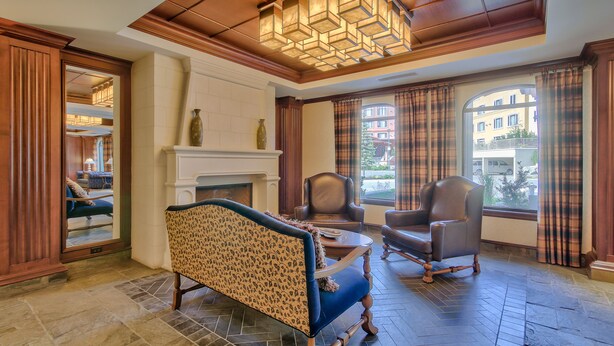 A spacious room with a fireplace, two leather chairs, a patterned sofa, and a view of buildings through large windows.