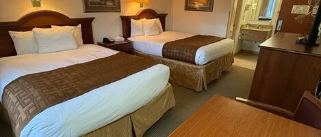 Une chambre d’hôtel avec deux lits, un bureau en bois, une chaise, une lampe de bureau, un miroir et une salle de bain visible à travers une porte ouverte.