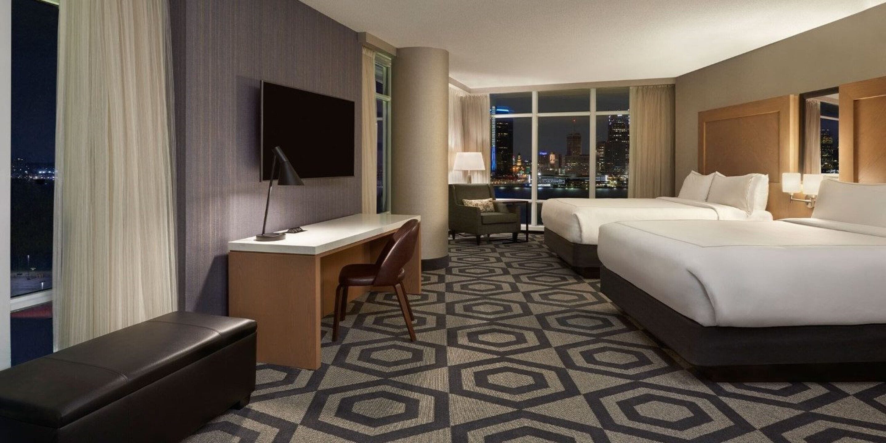Habitación de hotel con escritorio, dos camas, televisor y vista a la ciudad.