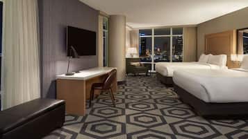 Habitación de hotel con escritorio, dos camas, televisor y vista a la ciudad.