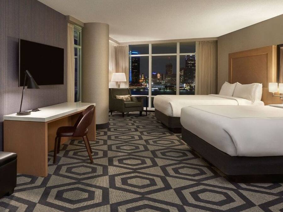 Habitación de hotel con escritorio, dos camas, televisor y vista a la ciudad.