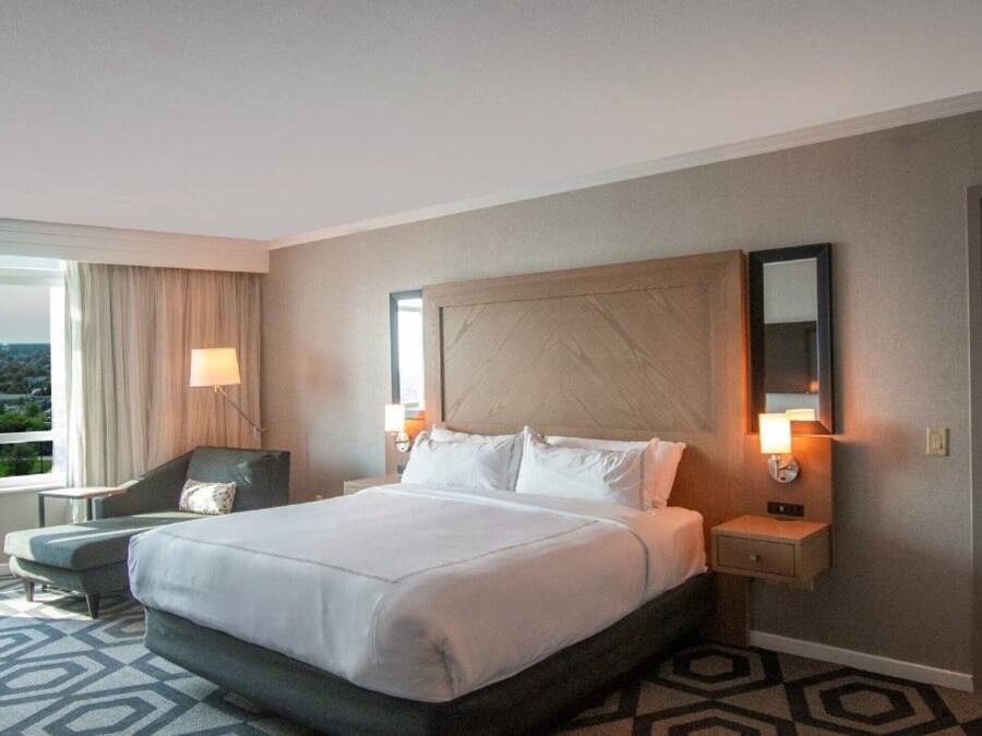 A hotel room with a large bed, a desk, a chair, a window with a view, and a patterned carpet.