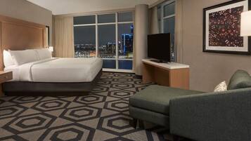 A hotel room with a large bed, a TV, a desk, and a view of the city at night.