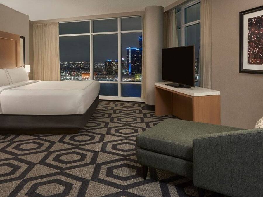 A hotel room with a large bed, a TV, a desk, and a view of the city at night.
