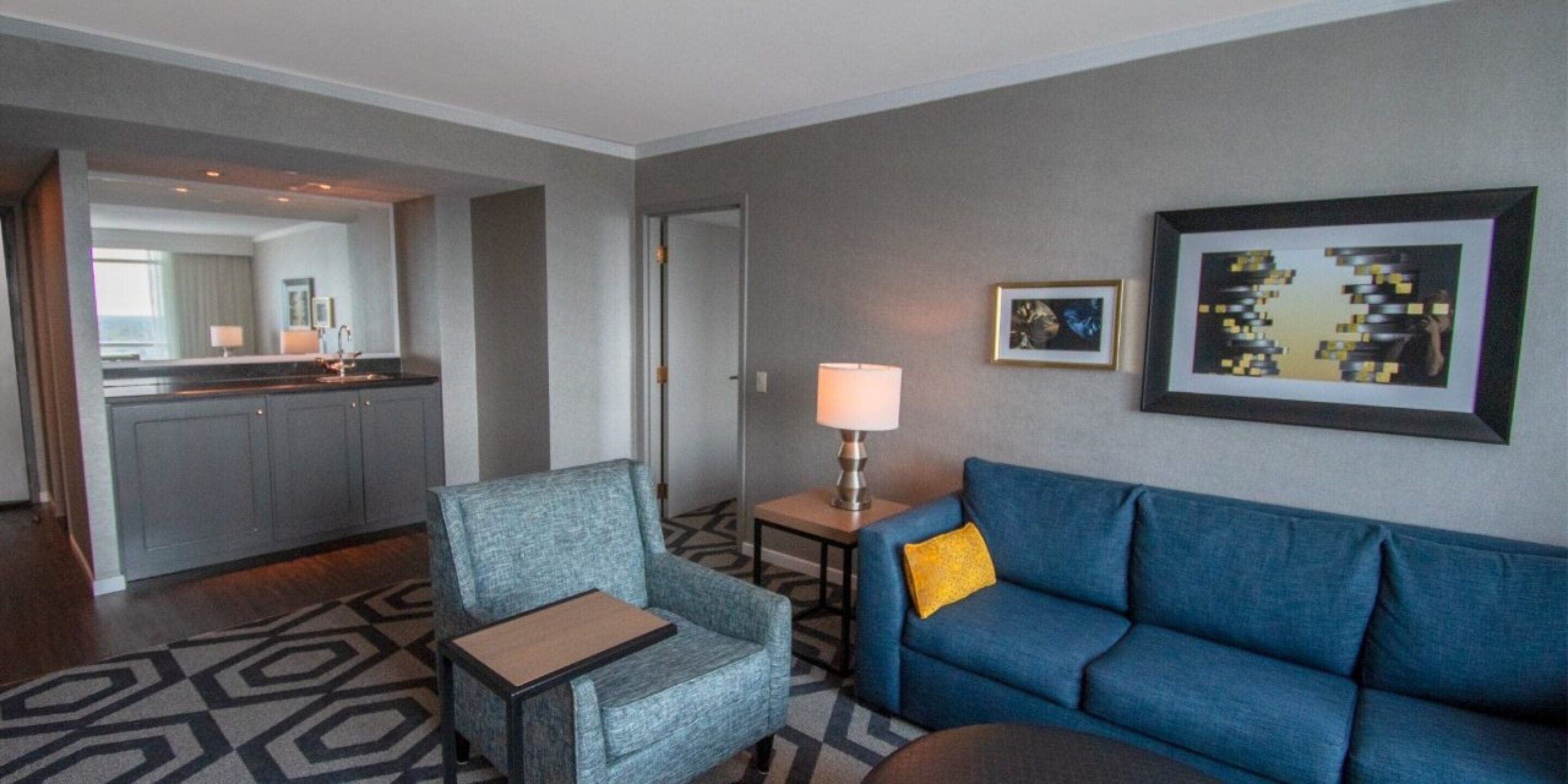 Una habitación de hotel con kitchenette, un sofá azul, un sillón gris y una alfombra estampada.