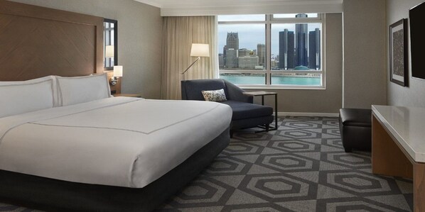A hotel room with a large bed, a desk, a chair, a view of the city, and a patterned carpet.