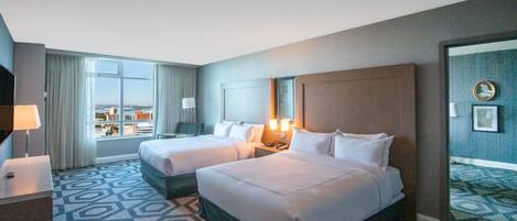 A hotel room with two beds, a large window with a city view, a patterned carpet, and a wall clock.