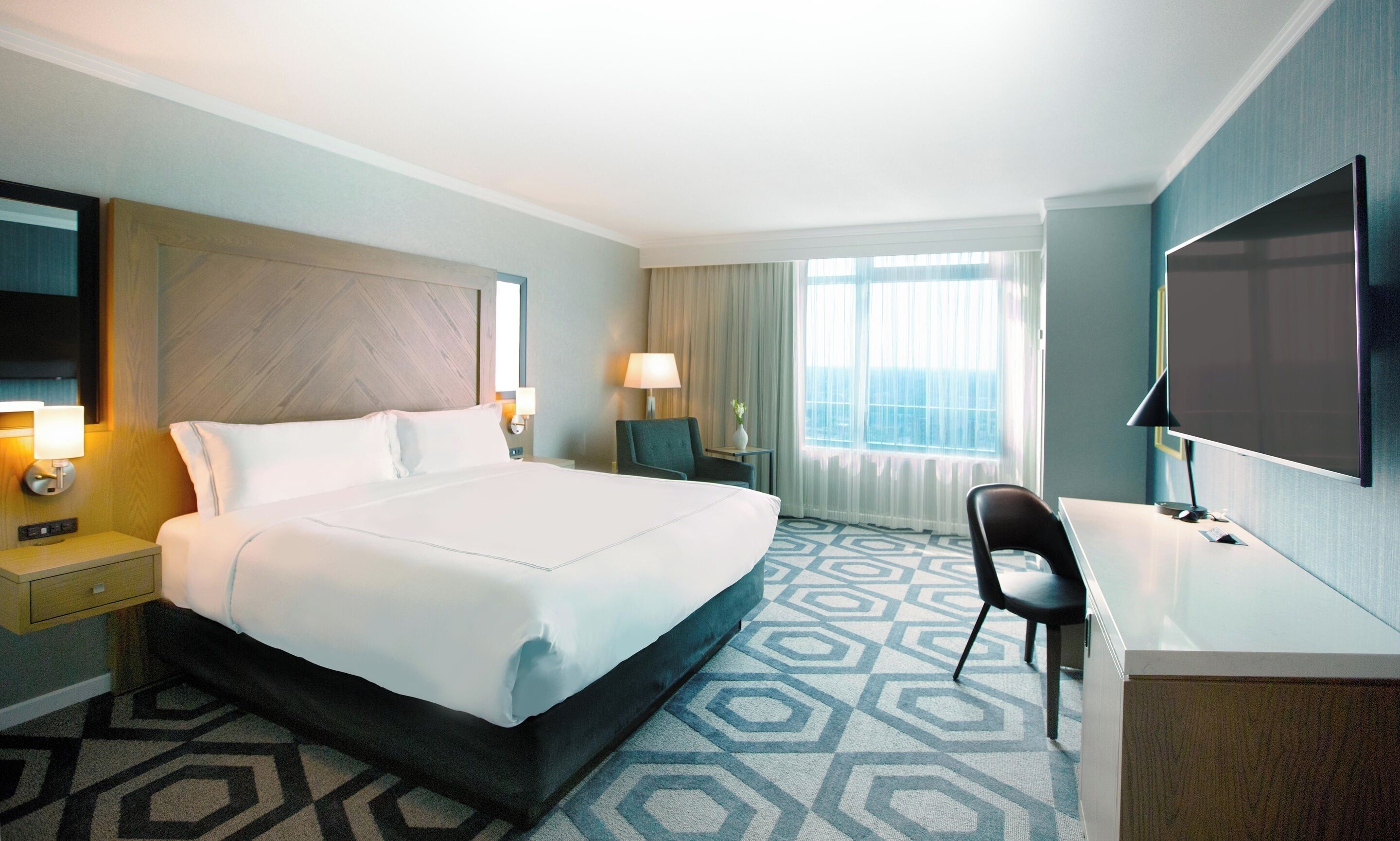 A hotel room with a bed, desk, chair, TV, lamp, and window.