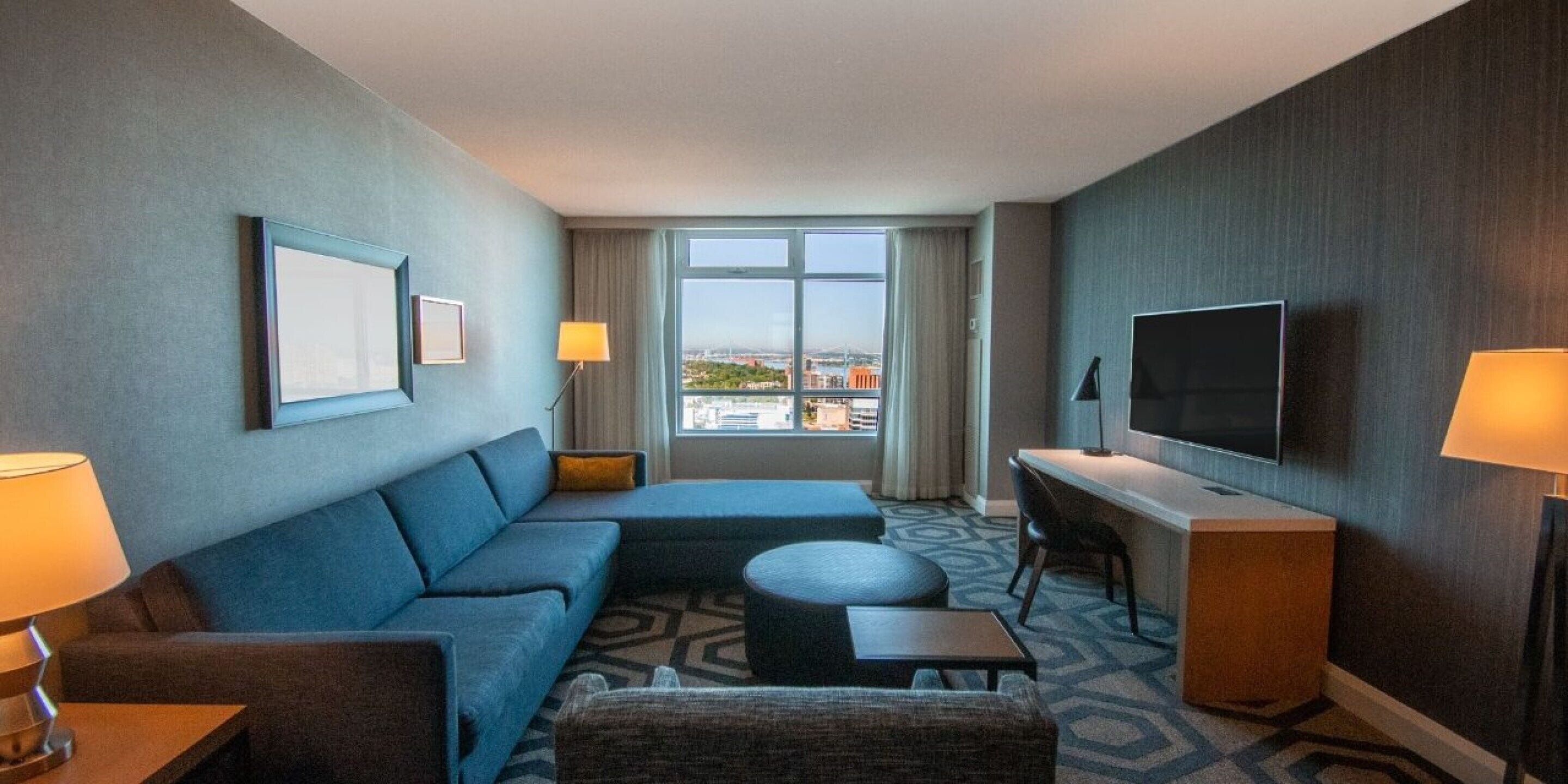 A hotel room with a sofa, a small table, a TV, and a window with a city view.
