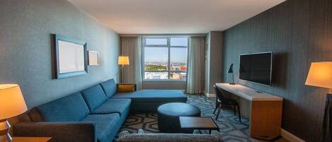 A hotel room with a sofa, a small table, a TV, and a window with a city view.