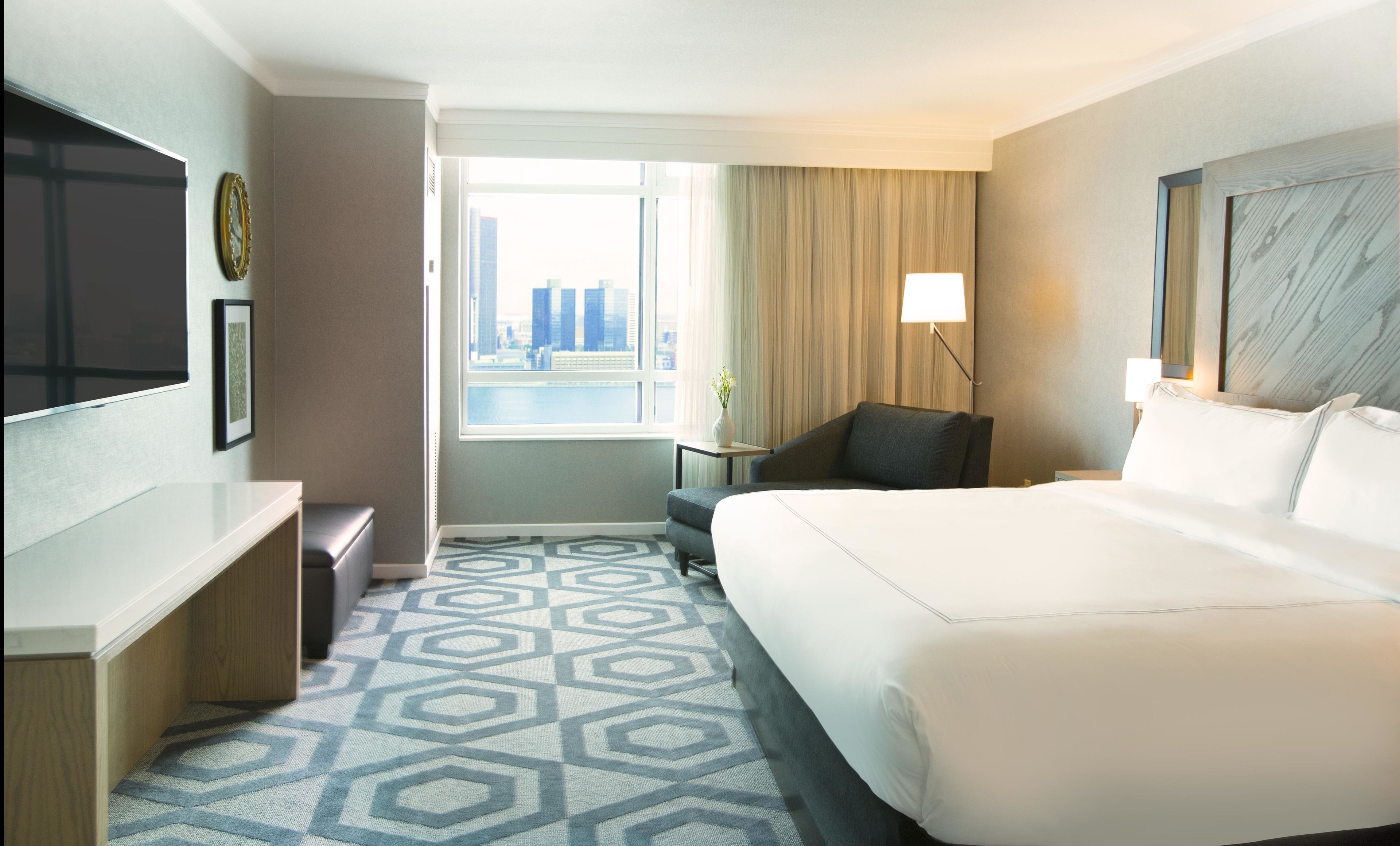 Habitación de hotel con una cama grande, un escritorio, una silla, un televisor, una ventana con vista a la ciudad y una alfombra estampada.