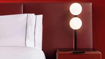 Uma cama bem arrumada com lençóis brancos, um painel de cama estofado em vermelho e uma mesinha de cabeceira com uma luminária moderna.