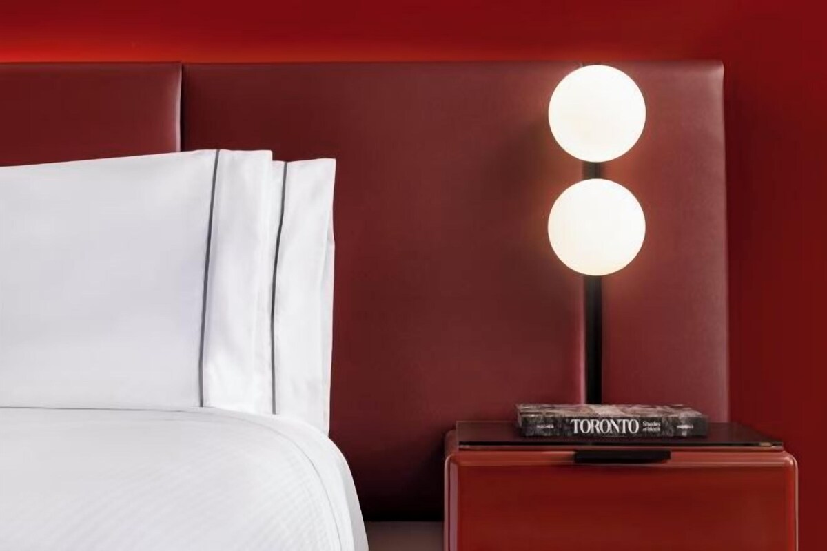 A neatly made bed with white linens and pillows, a red headboard, and a bedside table with a book titled "TORONTO" and a lamp.