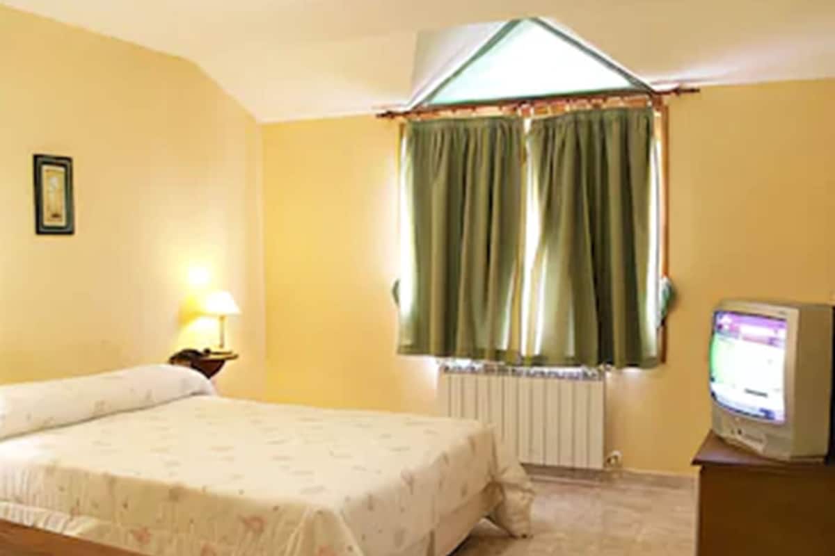 Una habitación con una cama, un escritorio, una lámpara, un ventanal con cortinas y un televisor.