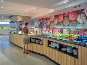 A buffet area with a variety of fruits and vegetables, a woman serving food, and a wall decorated with patterned umbrellas.