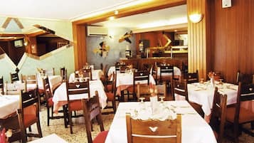 Ein Restaurant mit mehreren Tischen und Stühlen, ein Einblick in die Küche im Hintergrund sowie Holzwandverkleidung.