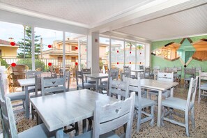 An indoor dining area with multiple tables and chairs, large windows, and a view of the outside buildings. - Villa Barbara (Santa Margalida)