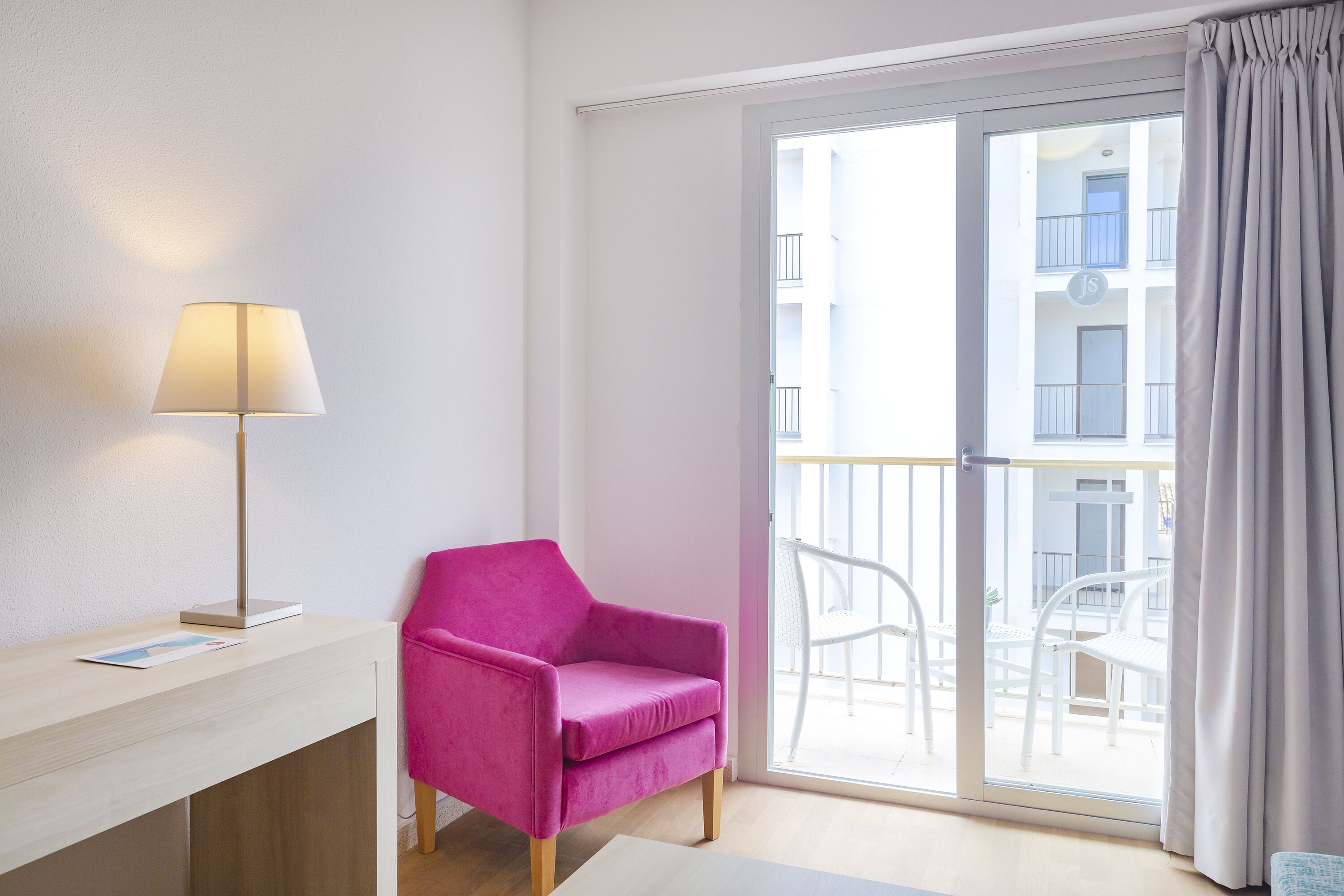 A room with a pink chair, a desk with a lamp, and a view of a balcony with white chairs.
