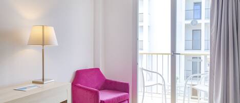 A room with a pink chair, a desk with a lamp, and a view of a balcony with white chairs.