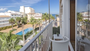 Un balcón con mobiliario de mimbre blanco, una piscina y vistas a la playa y edificios.