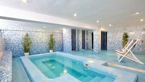 Una amplia piscina interior con mosaicos azules, rodeada de sillas de descanso y plantas en macetas.