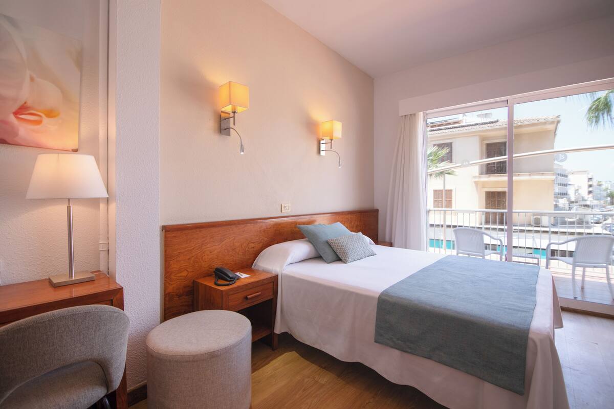 Ein Hotelzimmer mit Bett, Nachttisch, Lampe, Sessel und Blick auf den Pool sowie umliegende Gebäude.