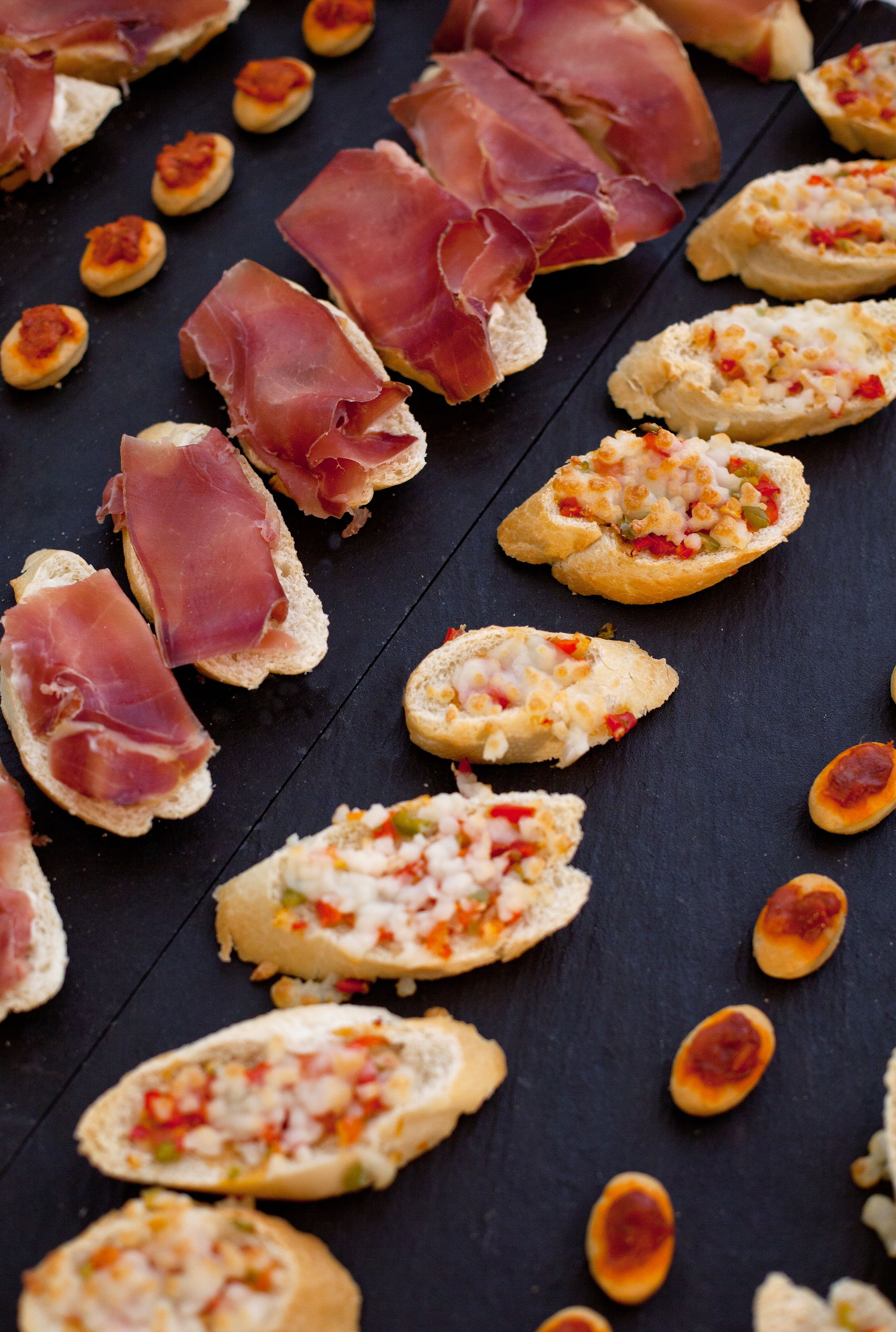 Une variété de hors-d'œuvre, notamment du pain accompagné de fromage et de poivrons rouges, et du pain garni de jambon de Parme, le tout présenté sur une surface sombre.