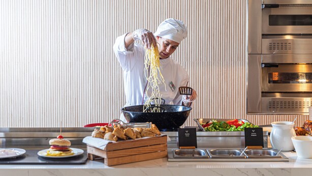 Un chef preparando comida en una cocina con un horno de acero inoxidable y una mesa de buffet con diferentes platos.