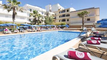 Una piscina con sillas de descanso y sombrillas, rodeada de edificios con balcones.