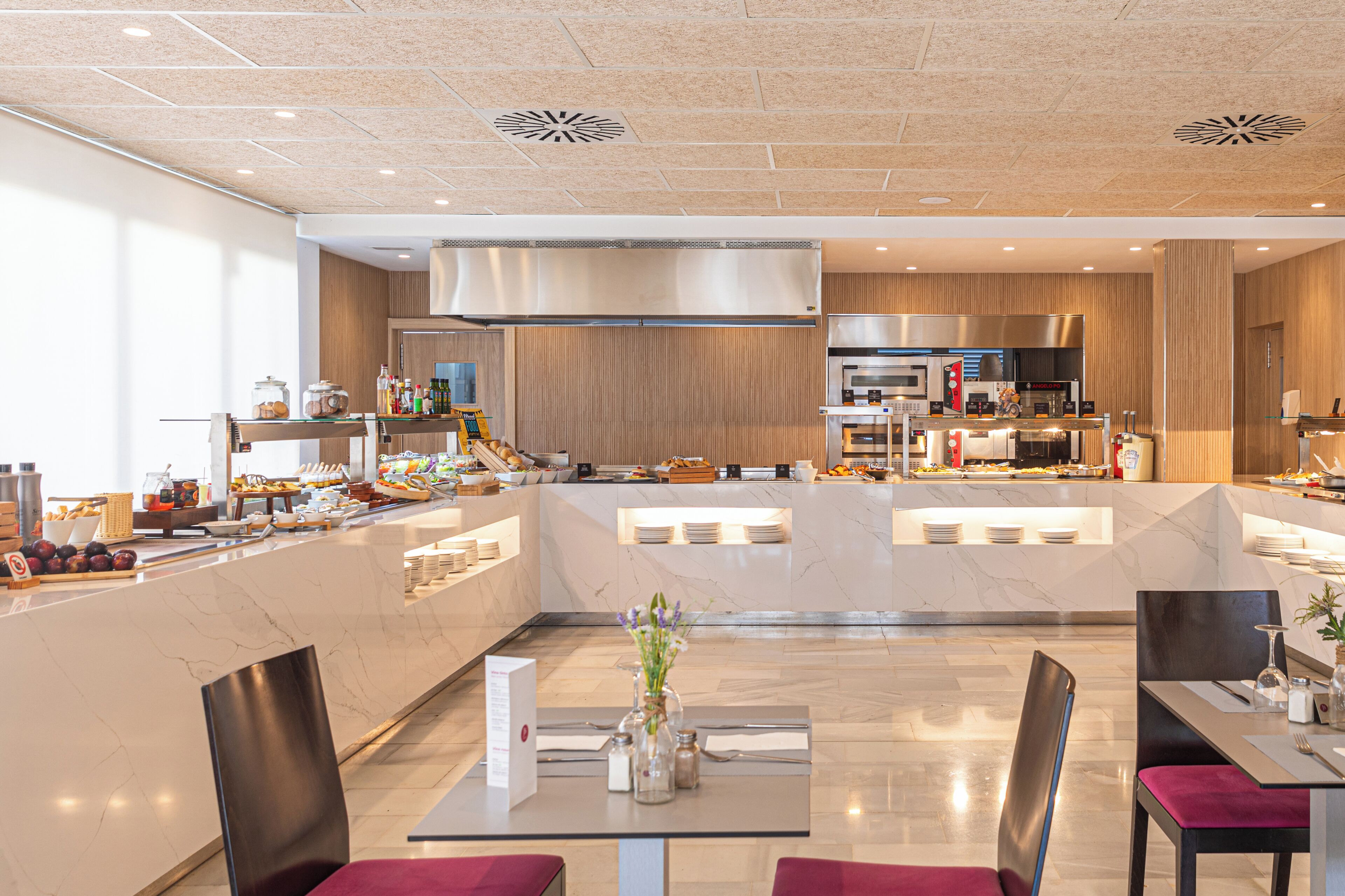 Un comedor moderno de hotel con una isla central, diferentes disposiciones de asientos y una cocina al fondo.