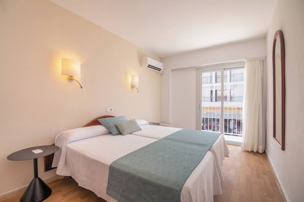 Ein Hotelzimmer mit zwei Betten, einem kleinen Tisch, einer Wand-Klimaanlage und einem Fenster mit Vorhängen.