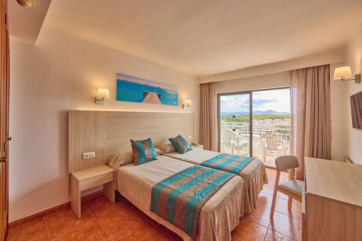 A hotel room with two beds, a desk, and a balcony with a view of the city.