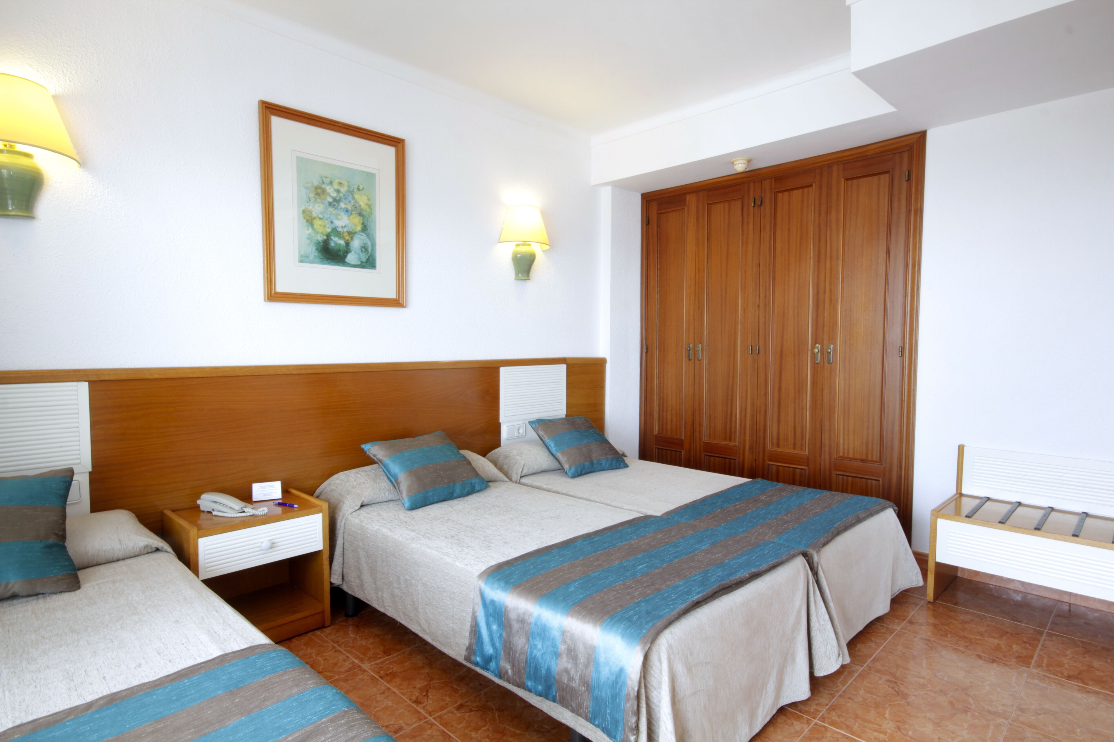 A hotel room with two beds, a wooden wardrobe, a nightstand with a lamp, and a framed picture on the wall.