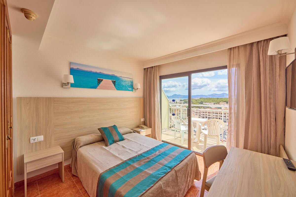 A hotel room with a bed, a desk, a chair, a nightstand, a lamp, a painting, and a view of the sea and mountains.