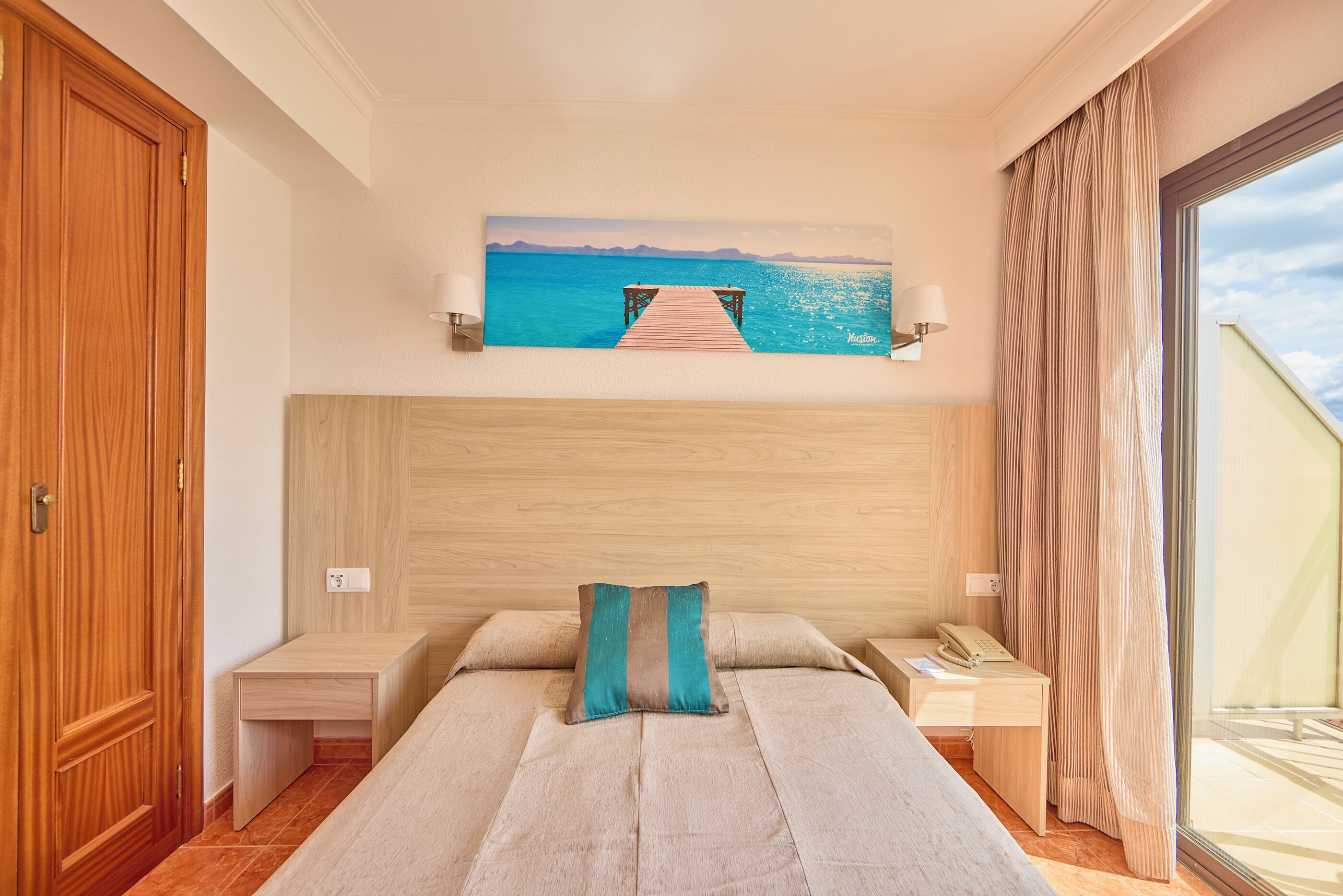 A hotel room with a bed, a wooden headboard, a painting of a pier, a bedside table, a nightstand, and a window with curtains.
