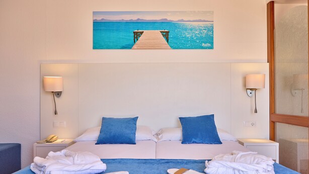 A double bed with blue and white bedding, two nightstands with lamps, a view of the sea through a window, and a painting of a pier. - Ilusion Markus & Spa (Santa Margalida)