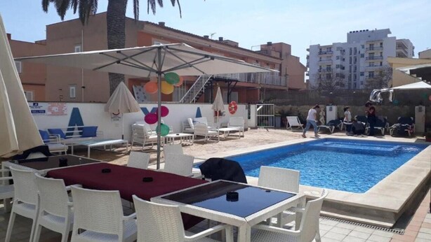 A poolside area with white lounge chairs, a table with a red tablecloth, and a covered seating area with umbrellas. - Raxa (Palma de Mallorca)
