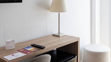 Een moderne hotelkamer met een houten bureau, een zwarte koelkast, een grijze stoel, een witte lamp en een aquarel kunstwerk aan de muur.