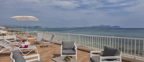 A balcony with white chairs and a table, overlooking a beach and the sea.