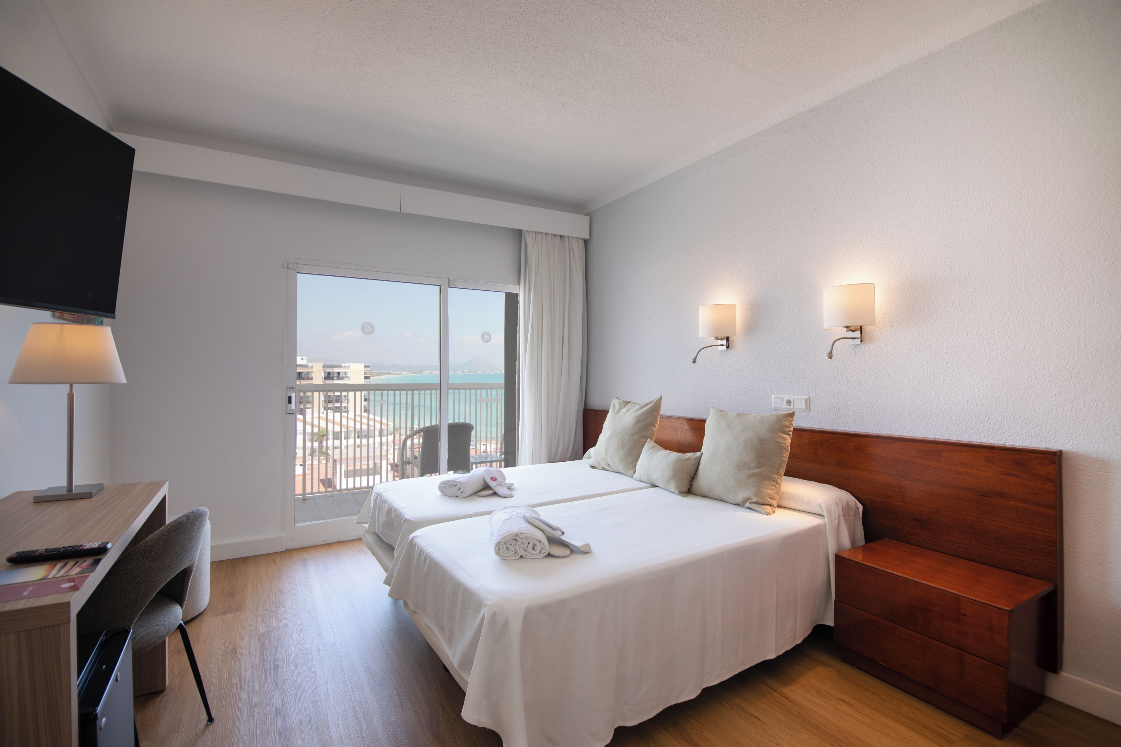 A hotel room with two beds, a desk, a chair, a TV, and a balcony with a view.