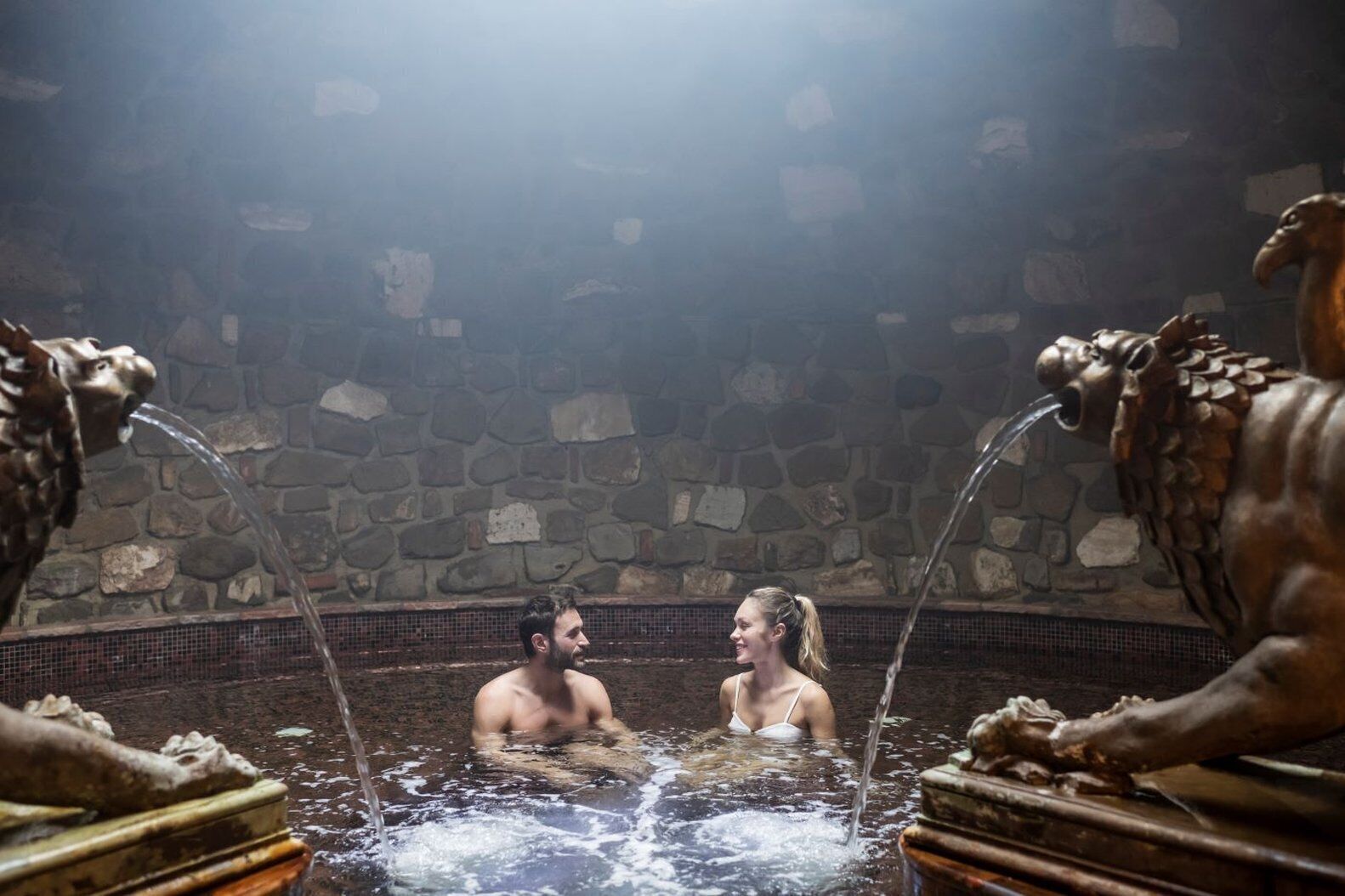 Ein Paar in einer Thermalquelle mit Steinsäulen, aus denen verziert gearbeitete Skulpturen Wasser speisen.