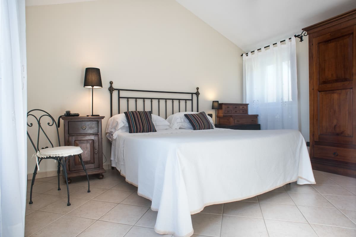 A bedroom with a bed, a nightstand, a chair, a wooden wardrobe, and a window with curtains.