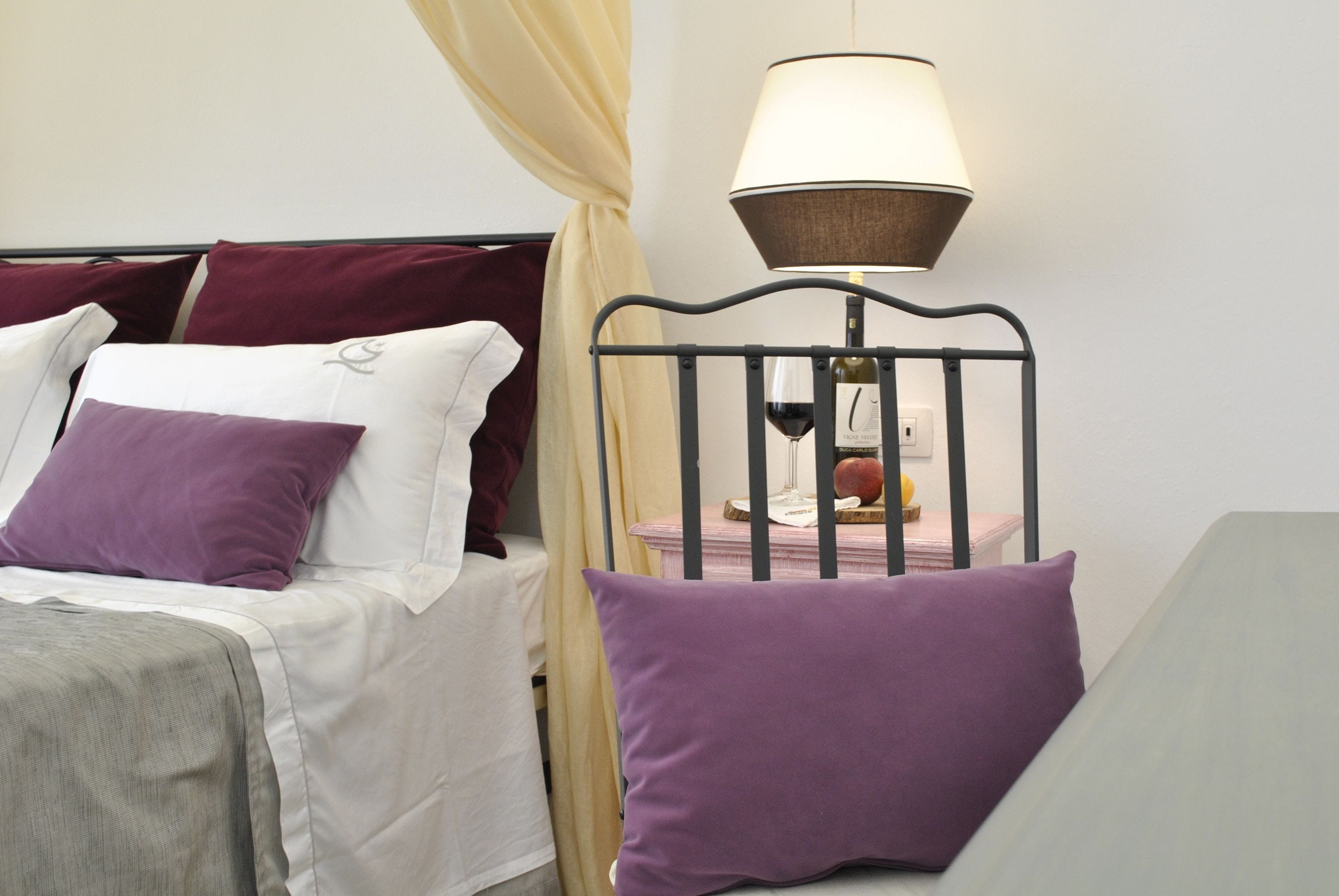 Una cama bien hecha con ropa de cama blanca y morada, un cabecero de metal negro y una mesita de noche con una lámpara y una botella de vino.
