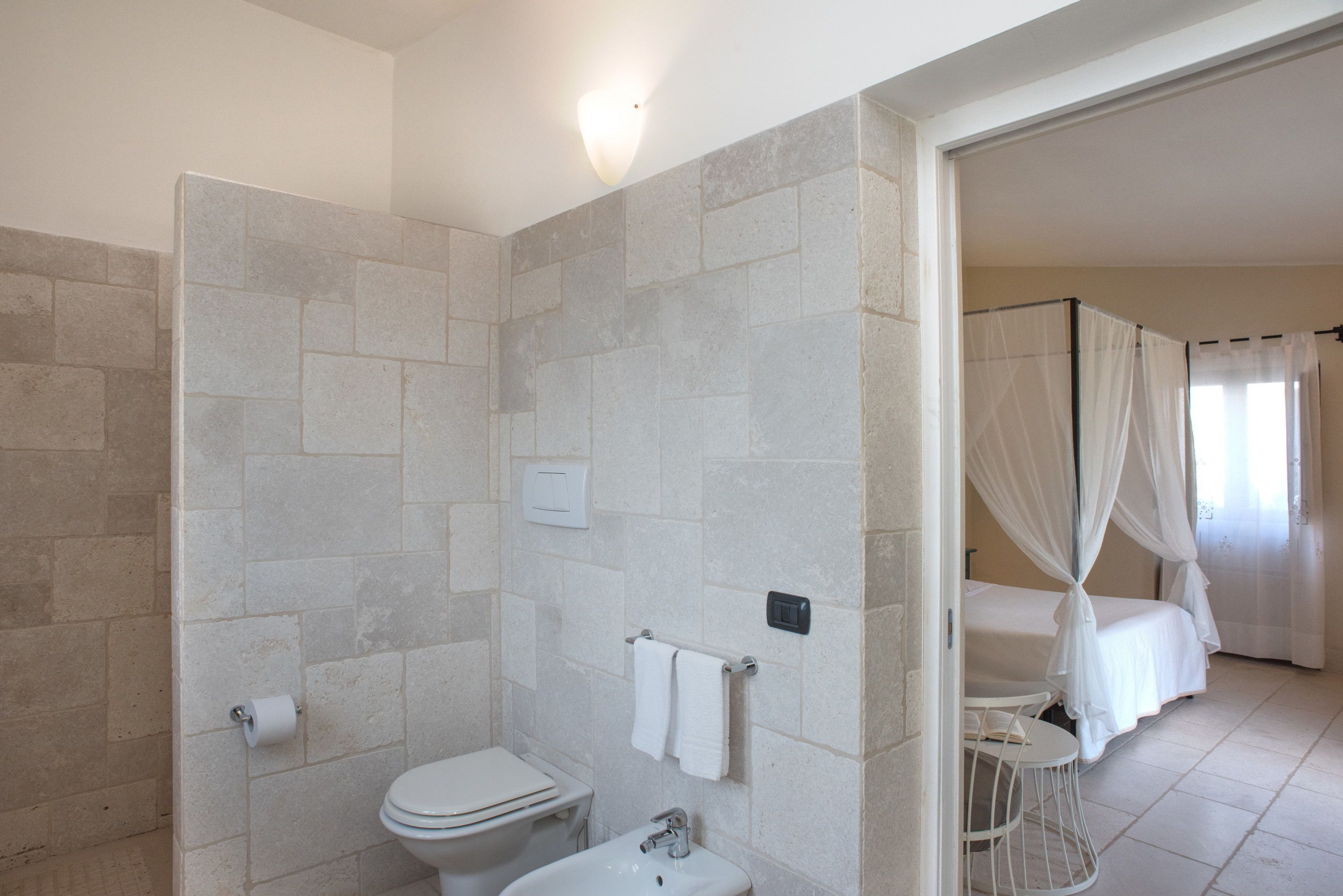Un baño con inodoro, lavamanos y zona de ducha. En la habitación contigua hay una cama con dosel.
