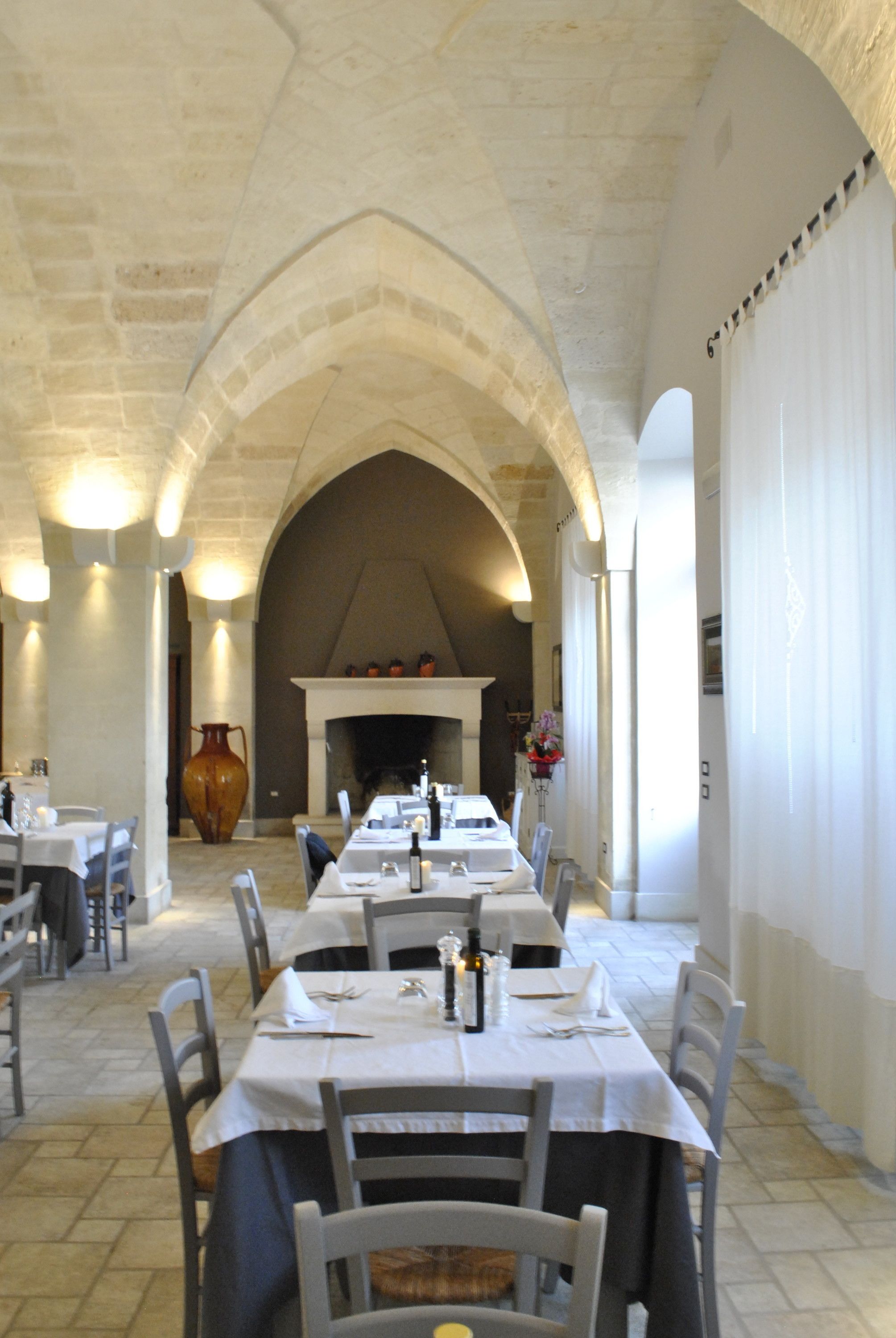 Interior com uma longa mesa de jantar posta para refeição, arcos de pedra e uma lareira ao fundo.