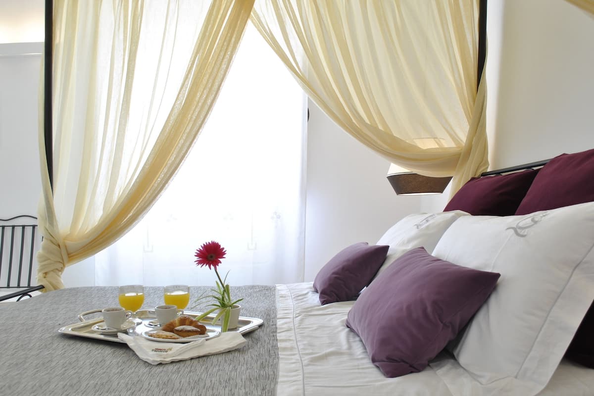 A neatly made bed with purple and white pillows, a tray with croissants, coffee cups, and orange juice, and a window with sheer curtains.