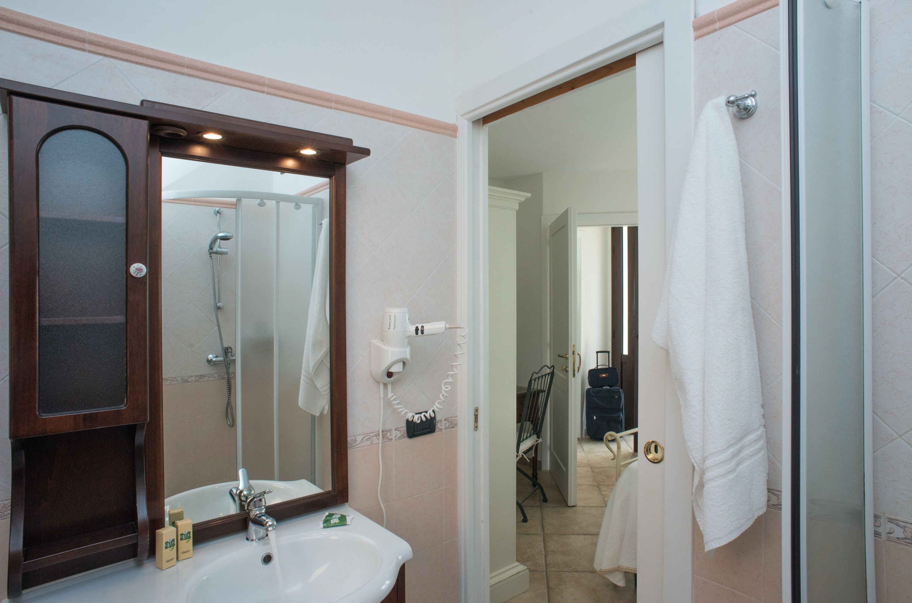 Baño con lavamanos, espejo y ducha. Hay una toalla colgada en la puerta y una maleta al fondo.
